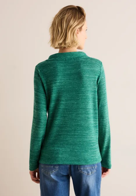Frau mit kurzem blondem Haar trägt grünen Pullover und Jeans, Rückenansicht.